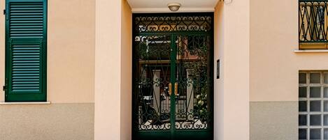 A building facade with a central entrance featuring a decorative metal gate, green shutters on windows, and a tiled pavement in front.