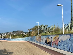 Ein gepflasterter Weg mit Wandgemälde, Straßenlaternen und Blick auf eine hügelige Stadt in der Ferne.
