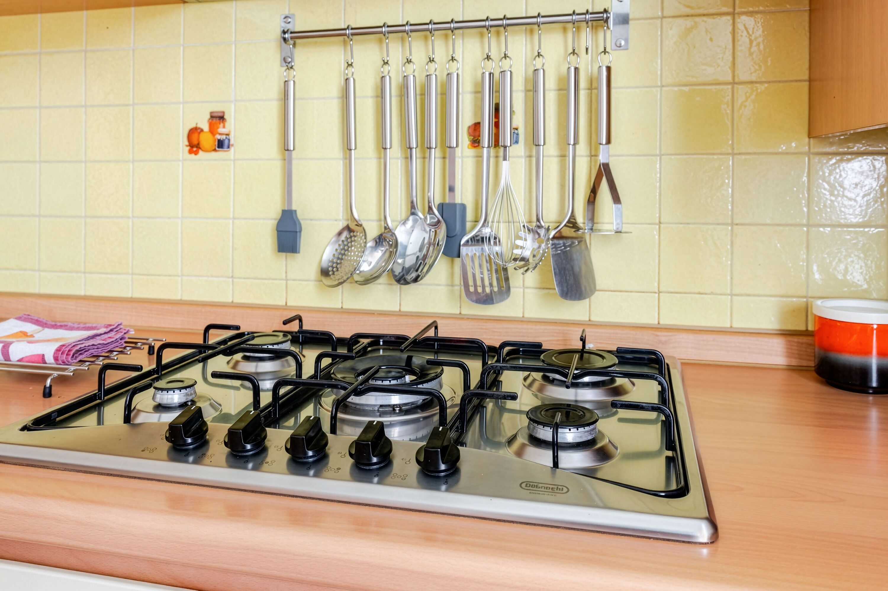 Een keuken met een gasfornuis, een hangende keukengerei-rek en een betegelde achterwand.