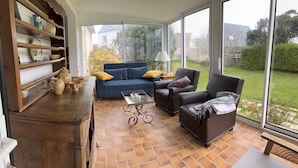 A sunlit room with a blue sofa, a brown armchair, and a glass coffee table. Shelves with decorative items line one wall. - the house of the end of the world (Porspoder)