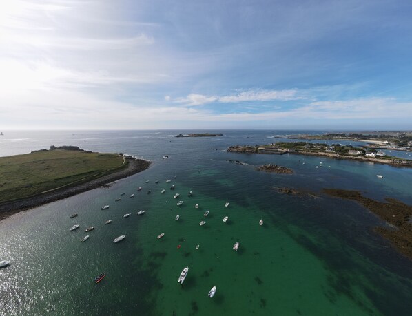 An aerial view of a coastal area with numerous boats anchored in clear waters, a rocky shoreline, and a distant view of a lighthouse. - the house of the end of the world (Porspoder)