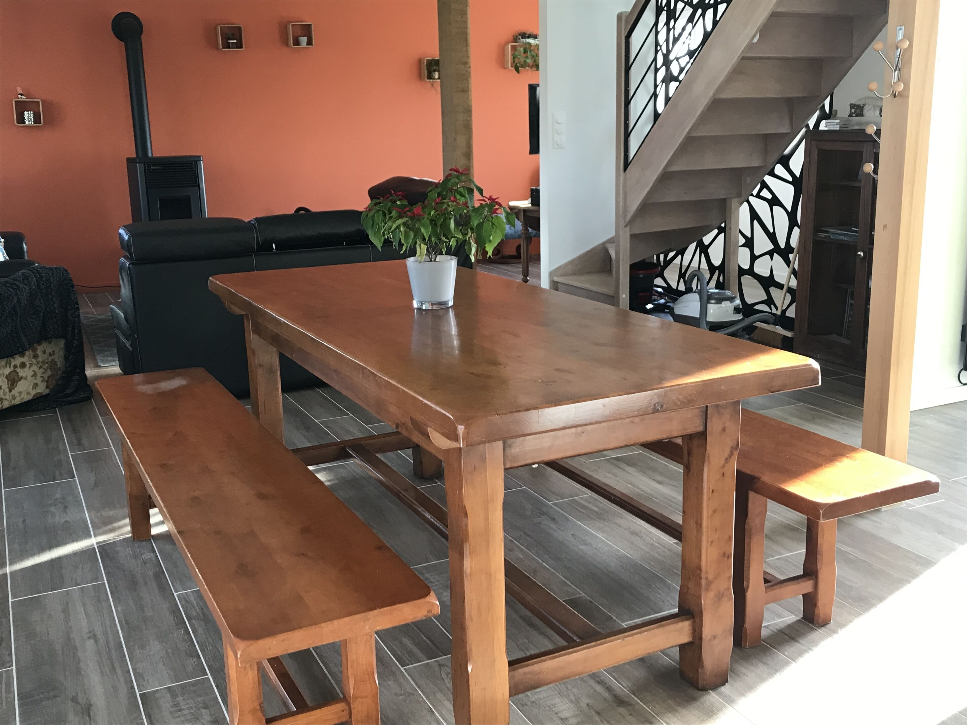 A wooden dining table with benches in a room with a tiled floor and a staircase in the background.