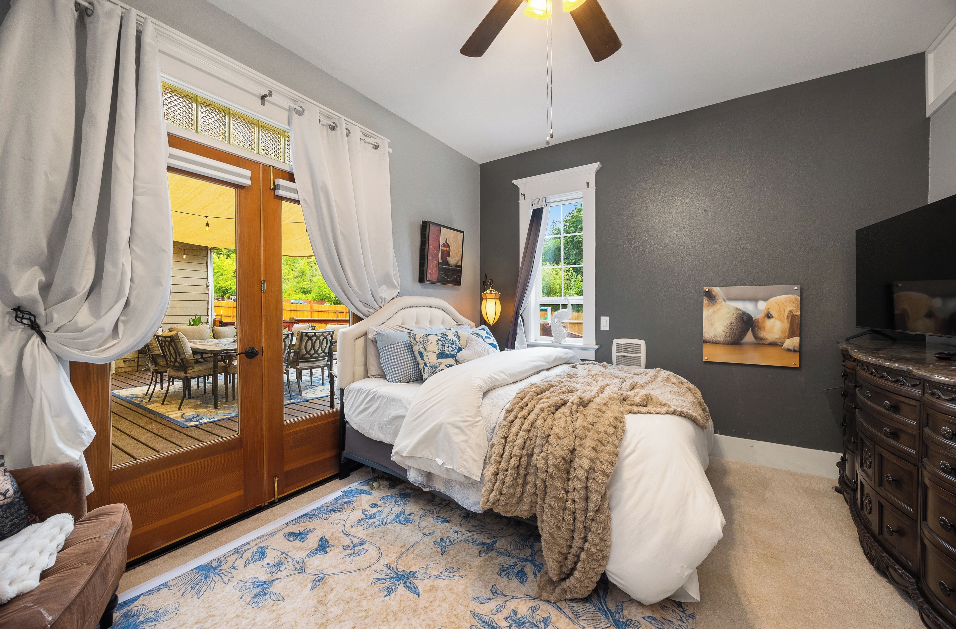 A bedroom with a bed, a dresser, a window with curtains, a ceiling fan, and a view of the outdoors.