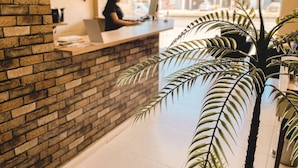 A reception desk with a brick wall, a woman working at a computer, and a large window with a view of the street. - Pousada São Pedro (Sao Pedro da Aldeia)