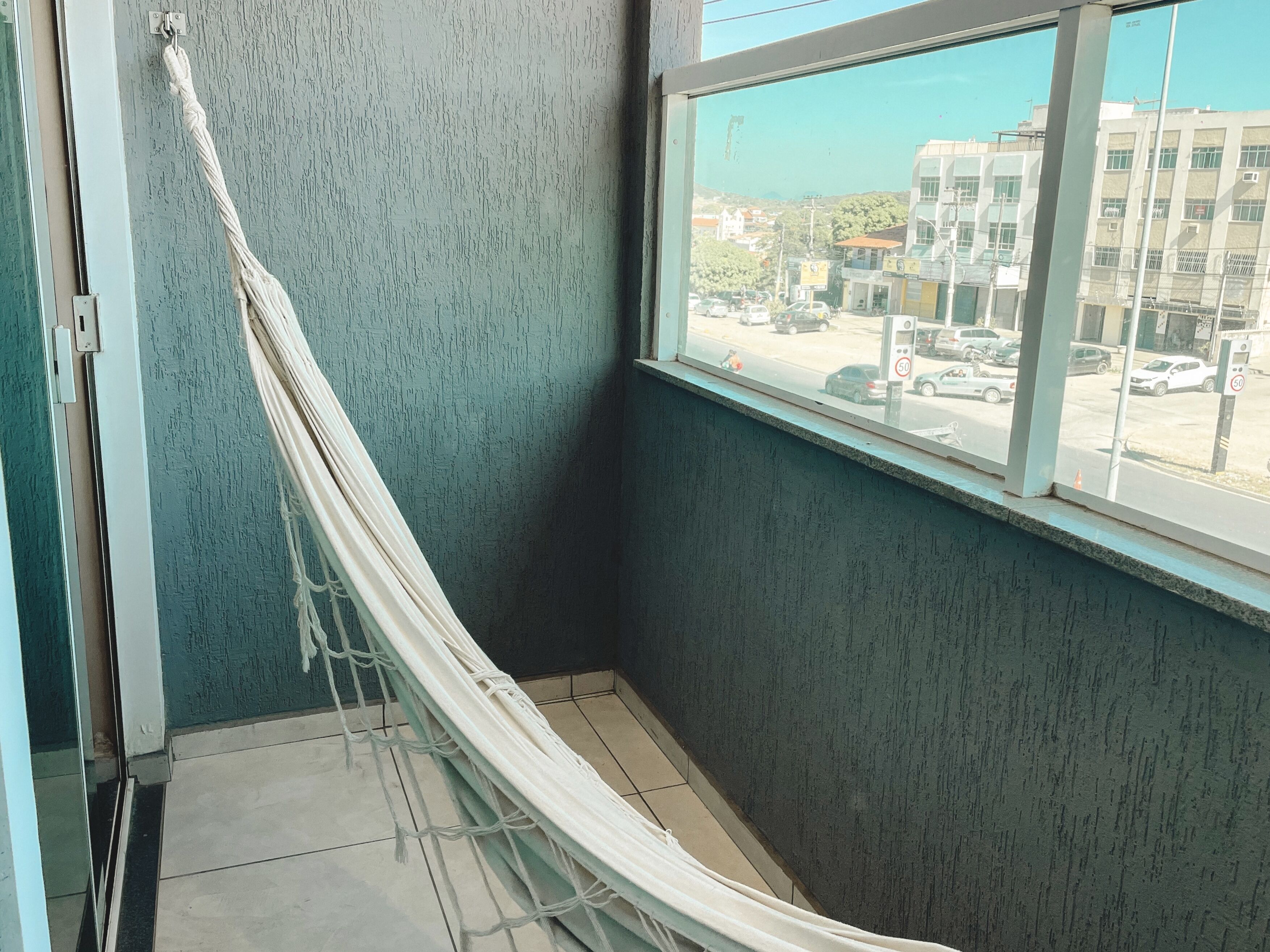 A room with a hammock against a textured teal wall, a window with a view of buildings and parked cars, and a tiled floor.
