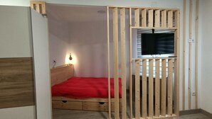 Quarto pequeno e fechado com uma cama de madeira, uma televisão presa a um painel de madeira e um cobertor vermelho sobre a cama.