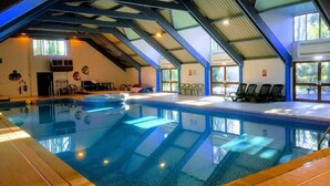 Una piscina interior con techos altos y tragaluces, rodeada de sillones de descanso y una pequeña mesa.