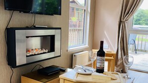 Una habitación moderna con televisor de pantalla plana, chimenea, una mesa de comedor con una botella de vino y copas, y vistas al exterior a través de una ventana.