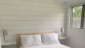 A neatly made bed with white linens and a blue throw blanket, a bedside table with a lamp, and a window with a view of greenery.
