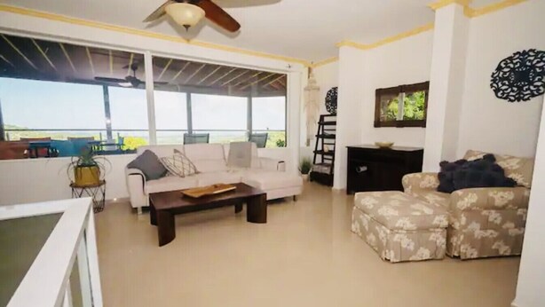 Ein geräumiges Wohnzimmer mit einem weißen Sofa, einem gemusterten Sessel und einem Couchtisch. Ein Deckenventilator, ein Fenster mit Blick und ein dekoratives Wandbild sind ebenfalls vorhanden.