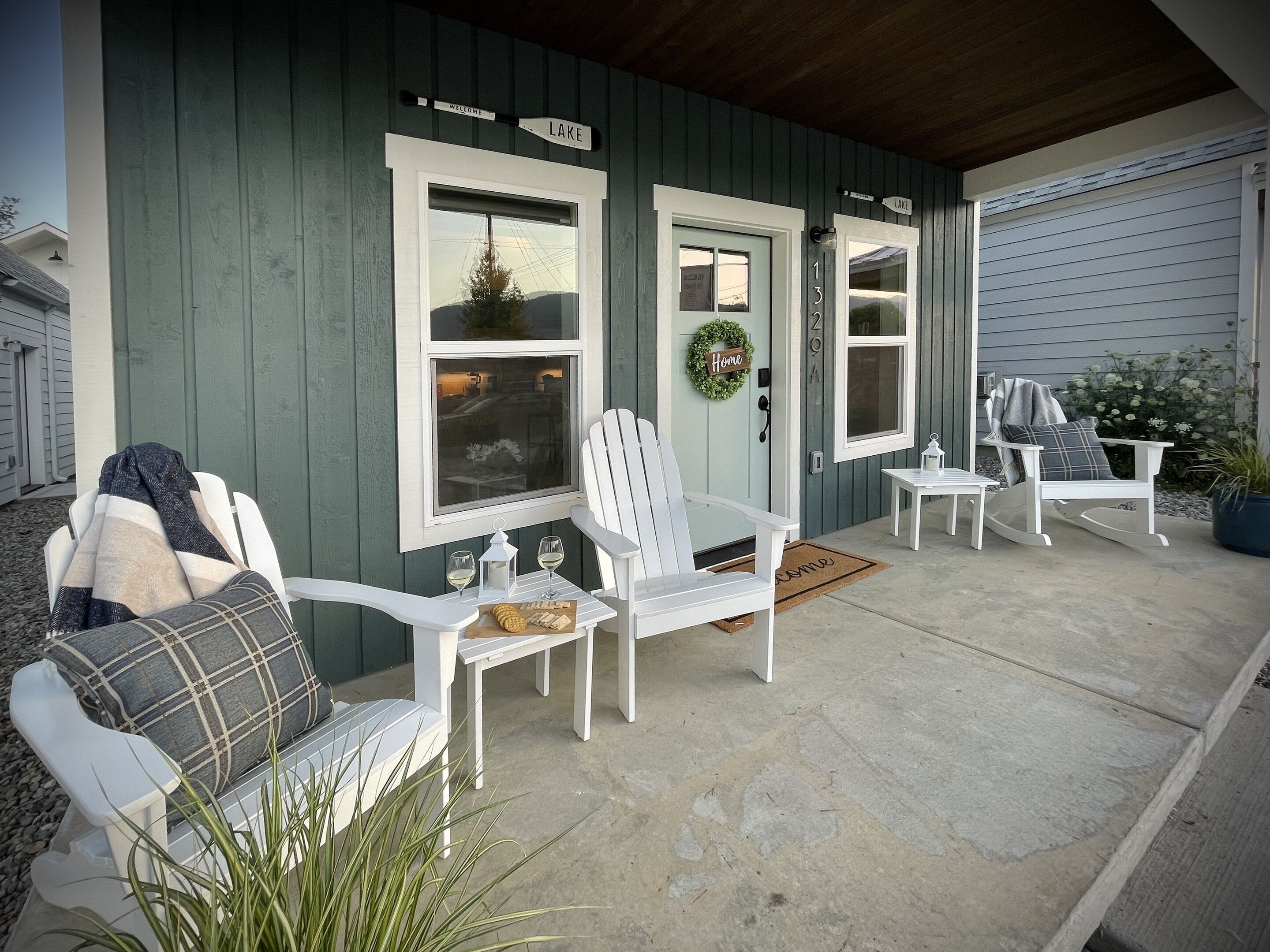A porch with white Adirondack chairs, a small table with a lantern, and a welcome sign.