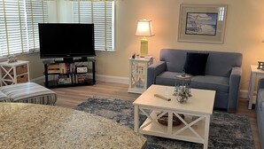 Un salon équipé d’un téléviseur à écran plat, d’un canapé gris, d’une table basse et d’une petite table blanche avec une lampe.