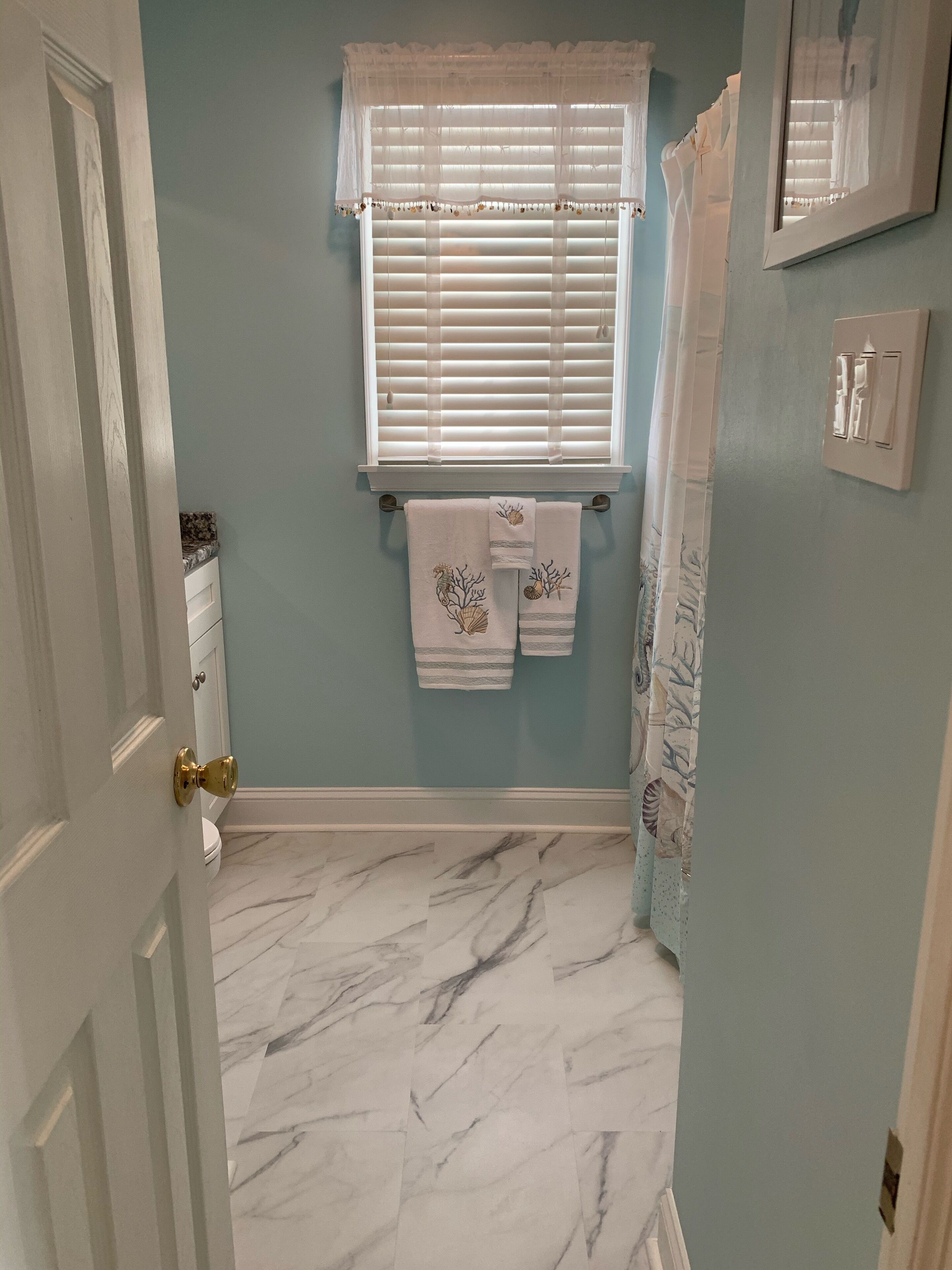 A bathroom with a marble floor, a window with blinds, a towel rack with two towels, and a shower curtain.
