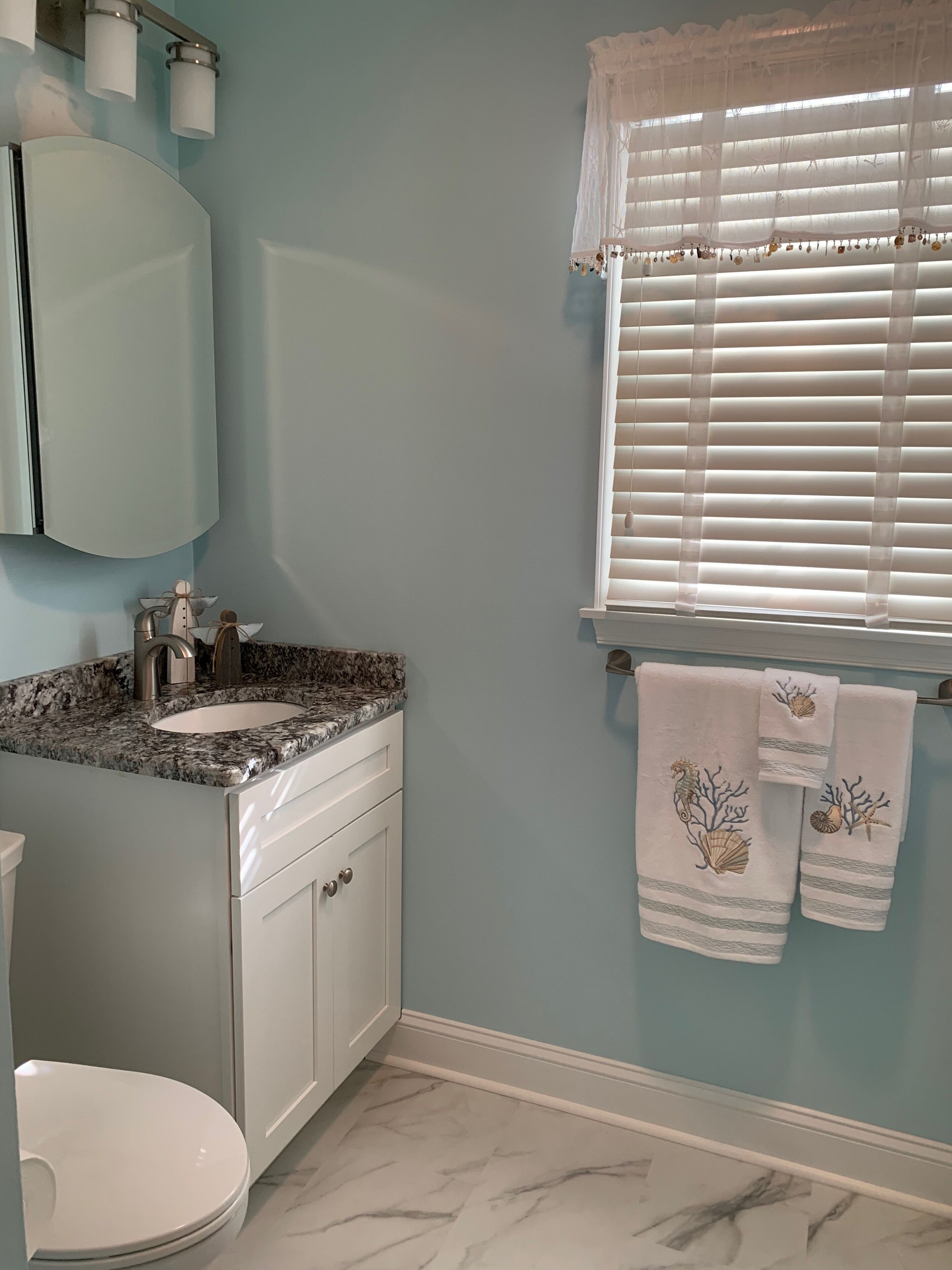 A bathroom with a marble sink, a white toilet, a mirror, and a window with blinds.