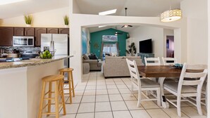 Eine moderne Küche mit einer zentralen Kochinsel, Barhocker, einem Essbereich mit Holztisch und weißen Stühlen sowie einem Wohnzimmer mit Sofa und Fernseher.