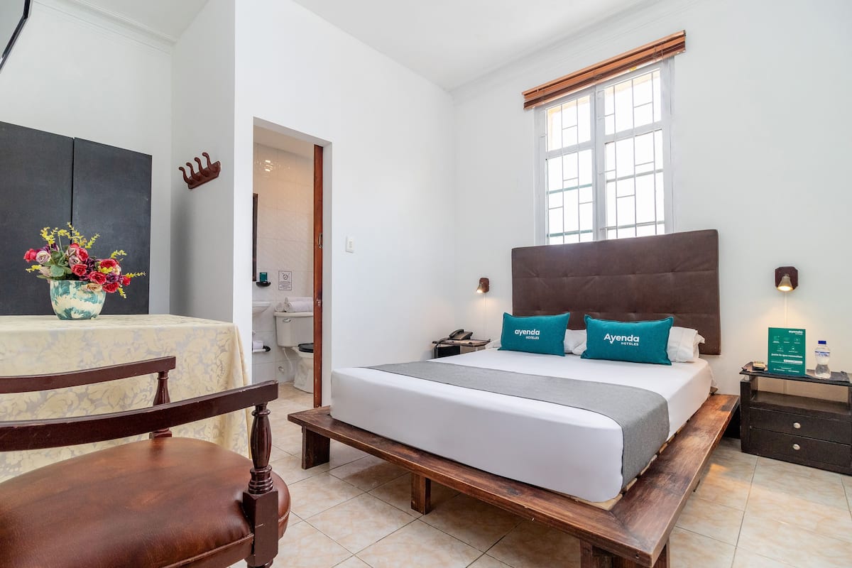 A hotel room with a bed, bedside tables, a chair, a desk, a vase with flowers, and a window with blinds.