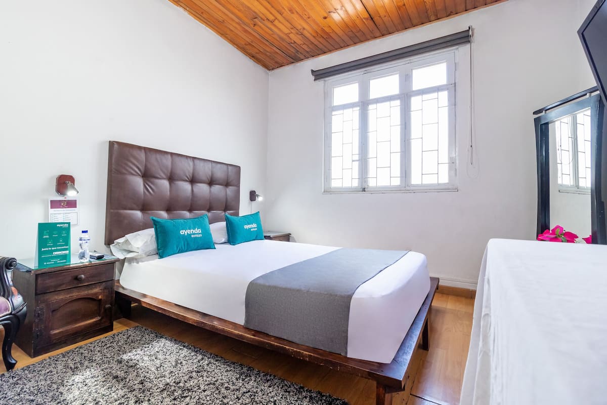 A bedroom with a large bed, wooden headboard, and a nightstand with a lamp. There is a window with blinds and a mirror on the wall.
