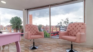 A room with two pink tufted chairs, a pink desk, and a large window with a view of buildings and a red bus. - Ayenda Eloina (Bogotá)