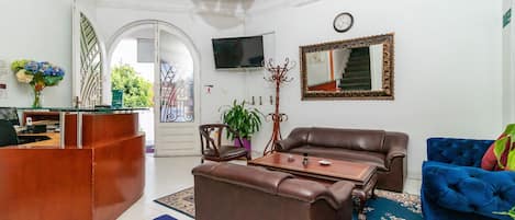 Una sala de estar con un sofá de cuero marrón, un sillón de terciopelo azul, una mesa de centro de madera y un televisor montado en la pared.