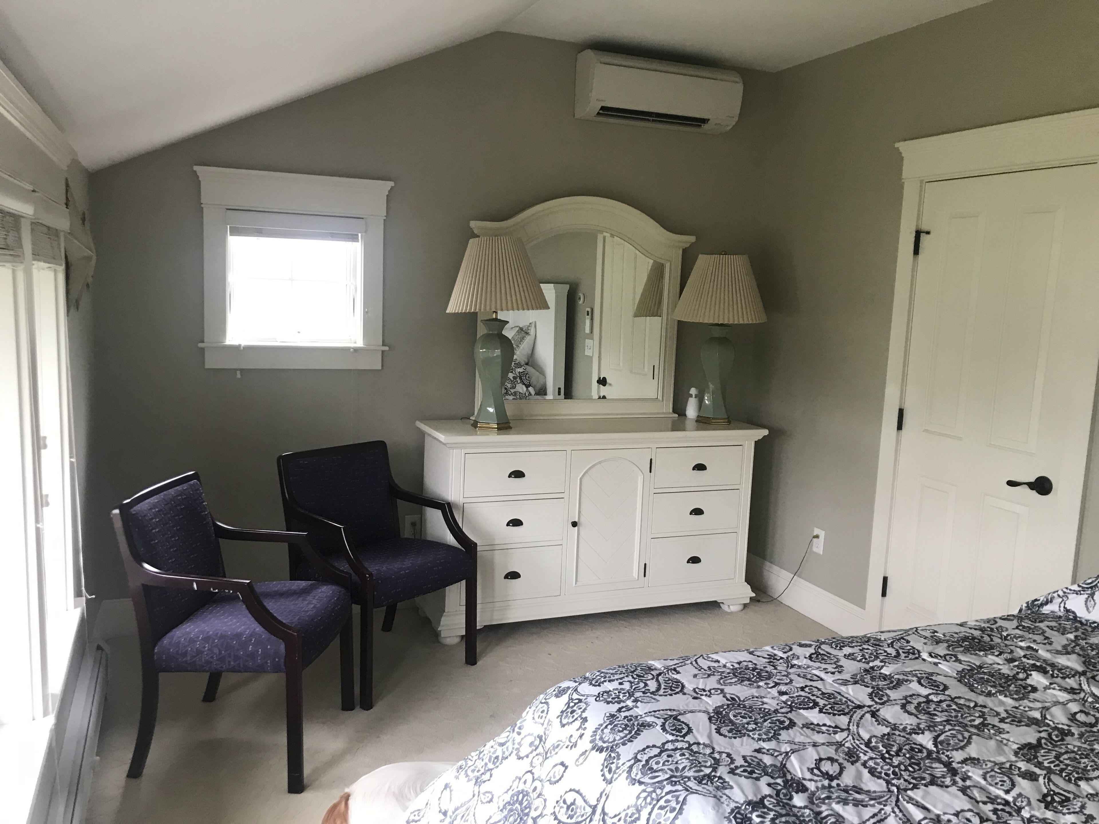 A bedroom with a white dresser, a chair, a bed with patterned bedding, a window, and an air conditioning unit.