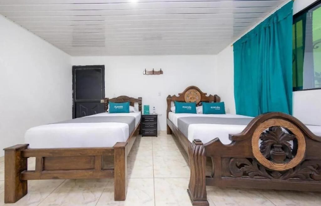 A room with two wooden beds, white bedding, teal pillows, a window with teal curtains, and a small table with a lamp.