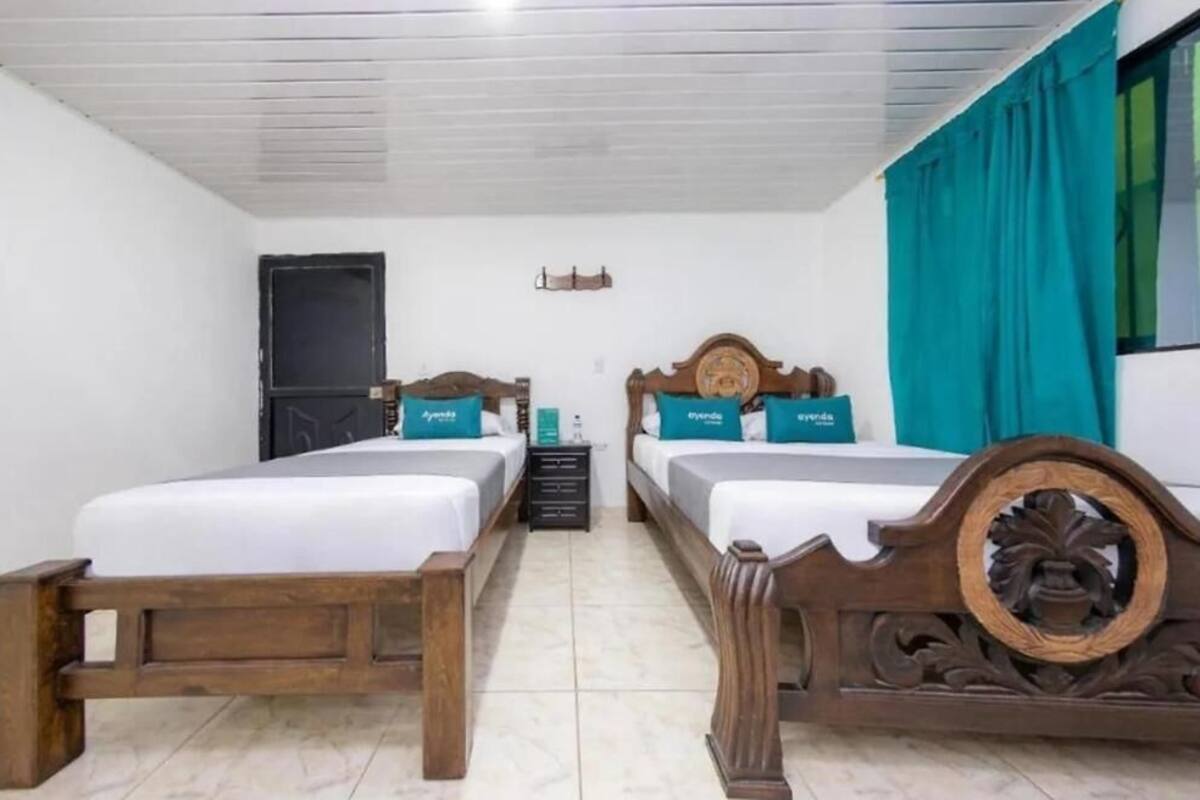 A room with two wooden beds, white bedding, teal pillows, a window with teal curtains, and a small table with a lamp.