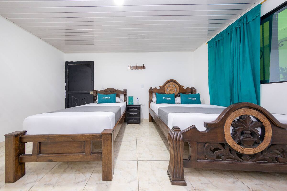 A hotel room with two wooden beds, white bedding, teal pillows, a wooden headboard, a small nightstand, and a window with teal curtains.