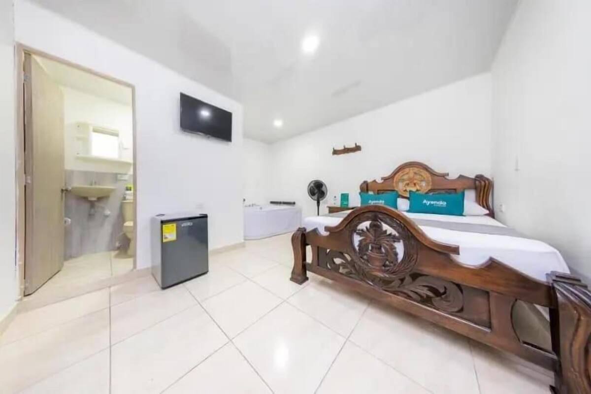 A bedroom with a wooden bed, a TV mounted on the wall, a small fridge, and a door leading to a bathroom.