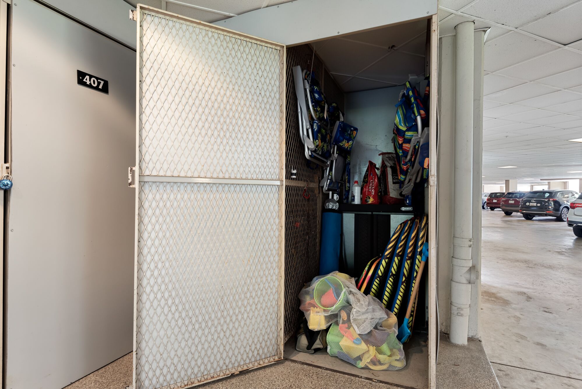 Un espace de rangement doté d’une porte en treillis marquée du numéro 407, contenant divers articles tels que des sacs, des cordes et une bouteille d’eau bleue.
