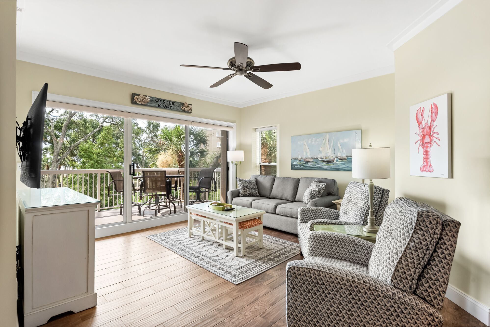 A living room with a sofa, chairs, a coffee table, a TV, a ceiling fan, a painting, and a balcony with a view of trees and a sailboat.