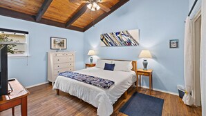 A bedroom with a large bed, wooden ceiling beams, a ceiling fan, two bedside tables with lamps, a dresser, and a window with blinds.
