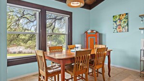 A dining area with a wooden table and chairs, a wicker pendant light, a colorful cabinet, and a view of trees through large windows.