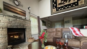 A cozy living room with a stone fireplace, a round wooden coffee table with a plate of cheese and wine glasses, a brown sofa, and a decorative wall hanging. - Sauna + Hot Tub | Mtn Views | Pet-Friendly Cabin (Gatlinburg)