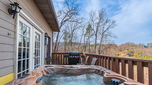 A hot tub on a wooden deck with a barbecue grill and chairs, overlooking a wooded area. - Sauna + Hot Tub | Mtn Views | Pet-Friendly Cabin (Gatlinburg)