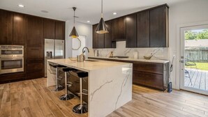 A modern kitchen with dark wood cabinets, a marble island with bar stools, stainless steel appliances, and pendant lighting. - Arthouse · Arthouse, Modern Luxury Getaway w/ Hot Tub near DT (Houston)