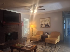 Un salon avec une cheminée, deux fauteuils, une table basse, une lampe et un ventilateur de plafond.