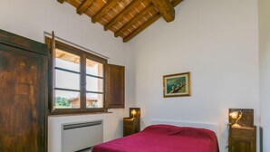 Un dormitorio con cama, armario de madera, ventana con contraventanas abiertas, radiador, un cuadro en la pared y lámparas de noche.