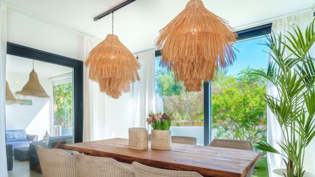Een eetgedeelte met een houten tafel, rieten stoelen en twee strooien plafondlampen.