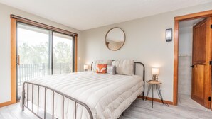Un dormitorio con cama, una mesita de noche con lámpara, un espejo en la pared y una puerta corredera de vidrio que da a un espacio exterior.