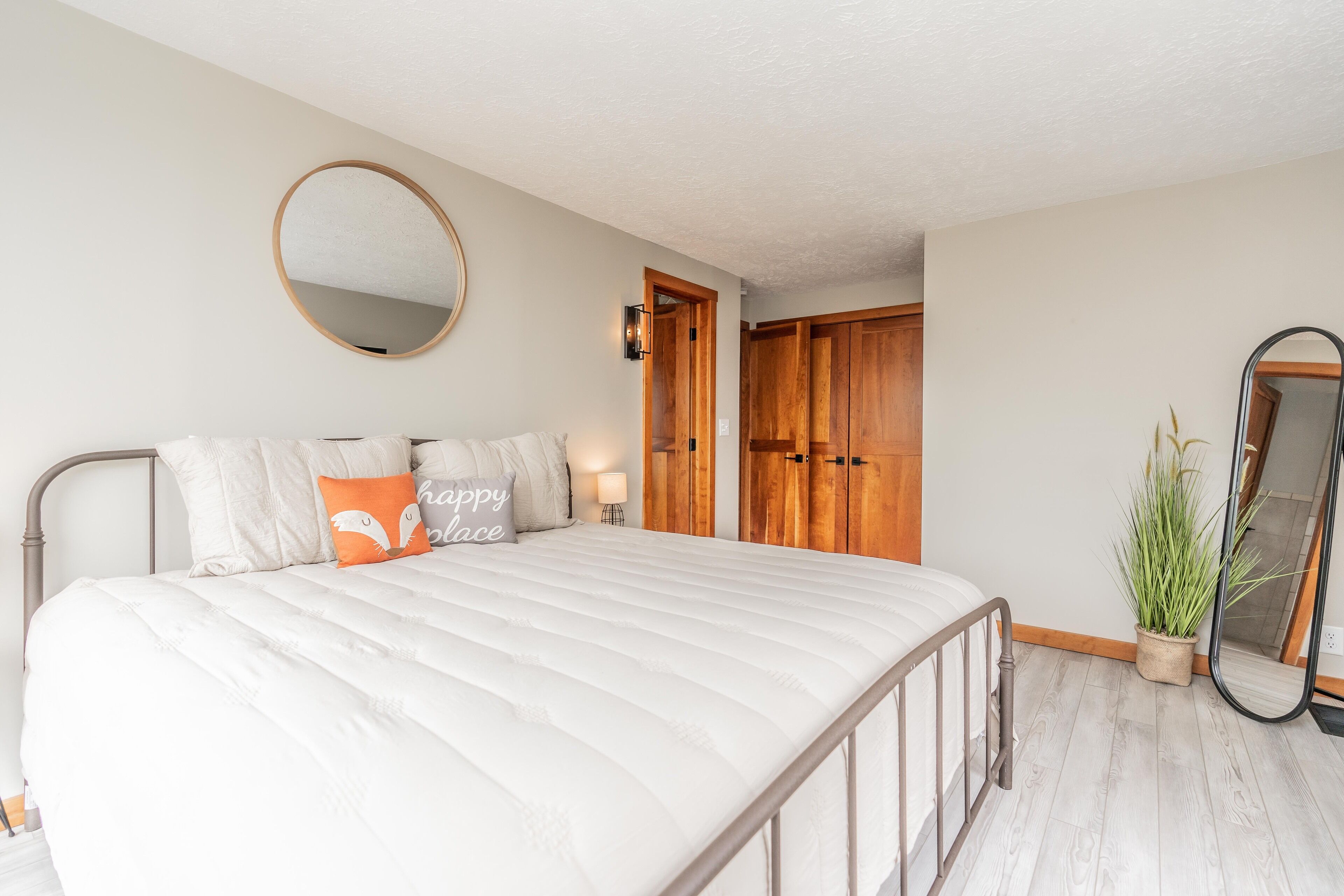 A bedroom with a large bed, a round mirror, wooden wardrobes, a bedside table with a lamp, and a potted plant.