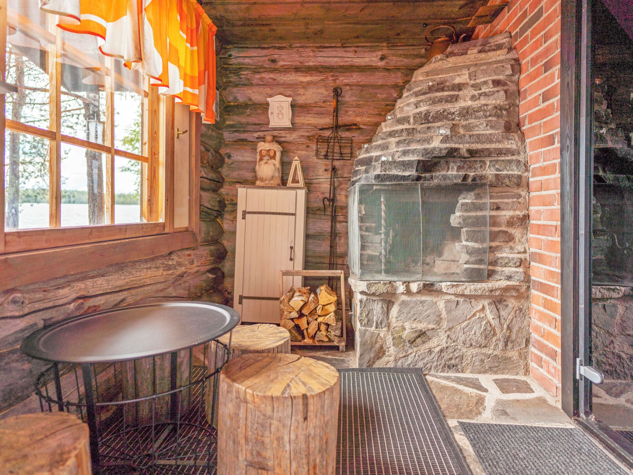 A rustic cabin interior with a stone fireplace, wooden walls, a metal table, and a window with curtains.