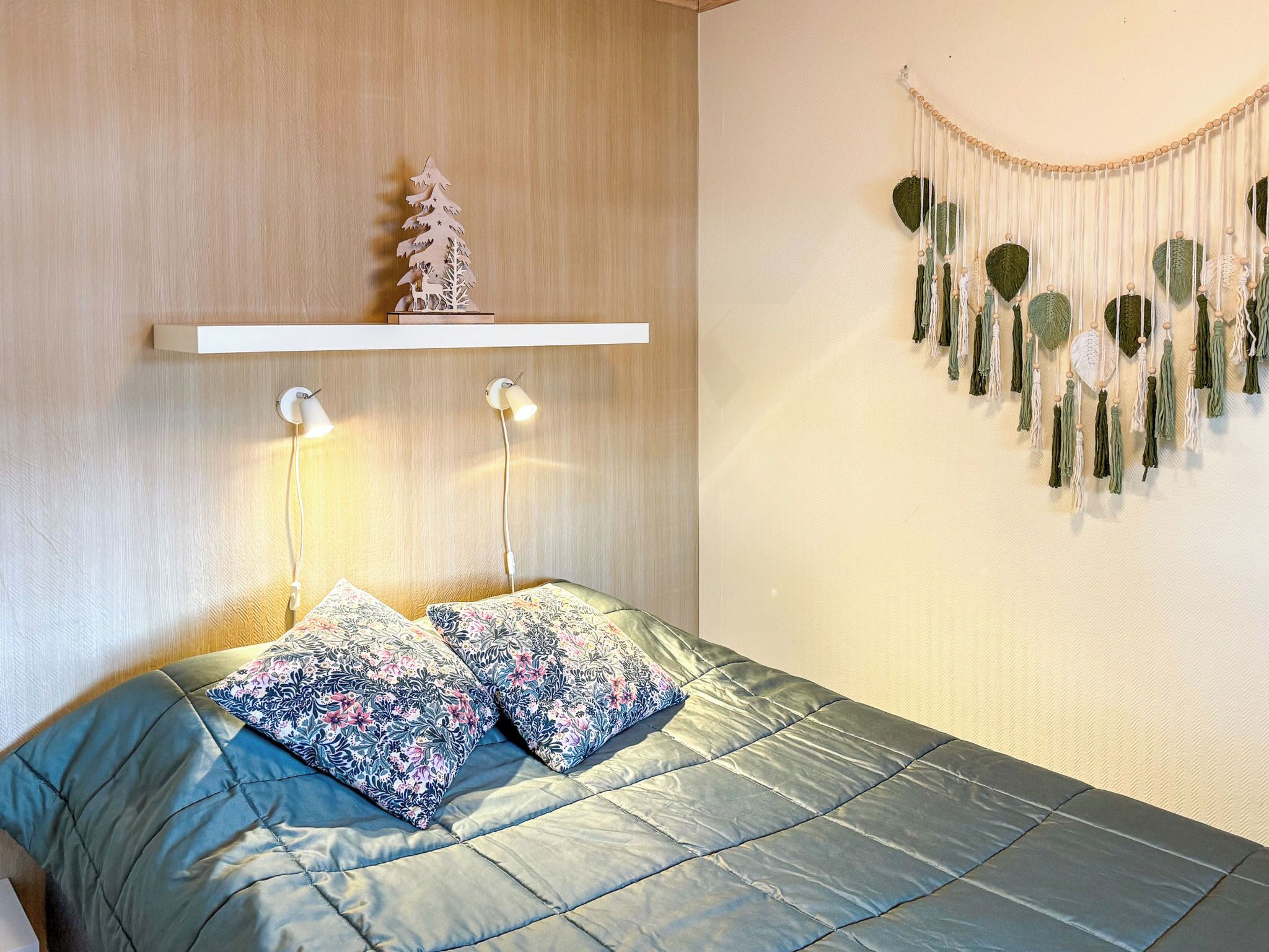 A bedroom with a bed, pillows, a wall-mounted shelf with a tree decoration, bedside lamps, and a wall hanging.
