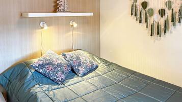 A bedroom with a bed, pillows, a wall-mounted shelf with a tree decoration, bedside lamps, and a wall hanging.