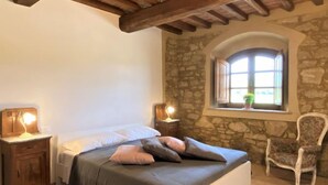 Une chambre avec un mur en pierre, une fenêtre donnant sur un beau paysage, un lit avec des oreillers, une chaise et des lampes de chevet.