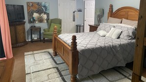 A bedroom with a wooden bed, a green armchair, a painting, and a television on a wooden cabinet. - Sea Trail Golf and Beach Get Way (Sunset Beach)