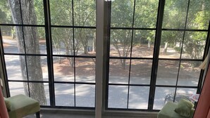 A room with a large window offering a view of a tree-lined street. There are two green armchairs and a small table with a blue floral tablecloth. - Sea Trail Golf and Beach Get Way (Sunset Beach)