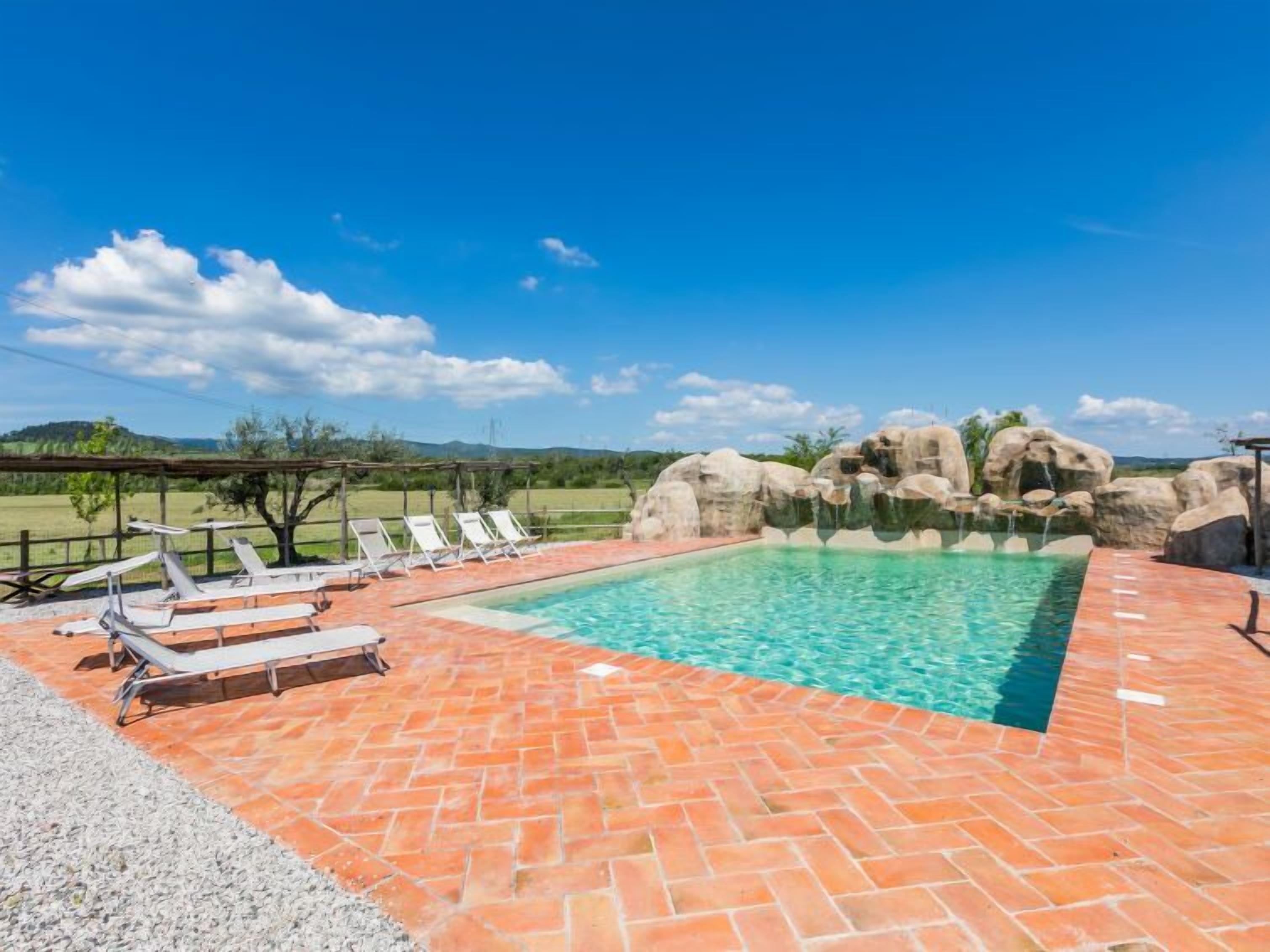 Ein rechteckiger Pool mit klarem, blauem Wasser, umgeben von einer Pflasterterrasse. Weiße Liegestühle und eine Pergola sind vorhanden.