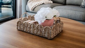 Habitació amb un sofà gris, una cistella de vimet amb flors blanques, una làmpada i un mirall gran a la paret.