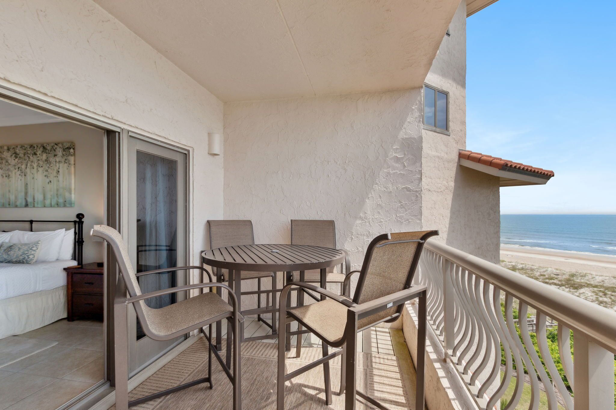 A balcony with a table and chairs, a bed, and a view of the beach.