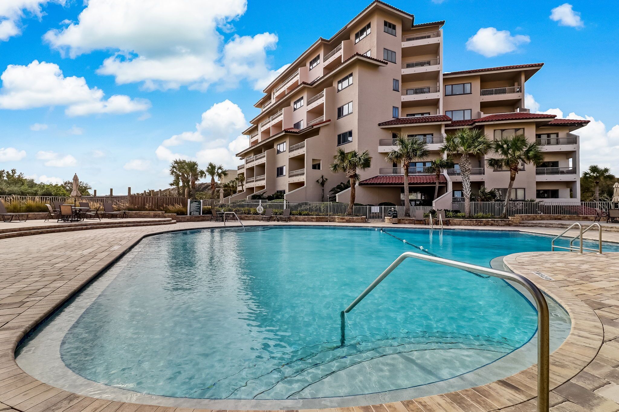 A swimming pool with a curved edge, surrounded by a wooden deck. In the background, there is a multi-story building with balconies and palm trees.